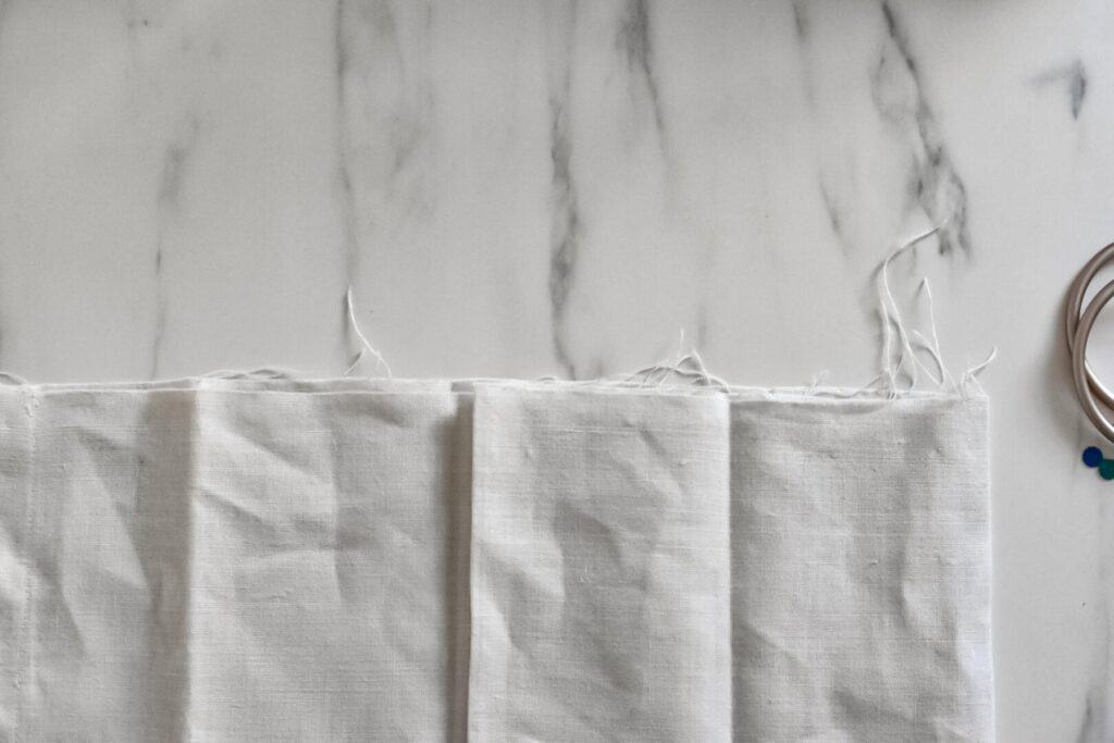 Beige fabric with frayed edges is laid out on a marble surface, showing several vertical folds, ready to make a DIY linen ring sling. A metal sling ring is partially visible on the right side of the image.