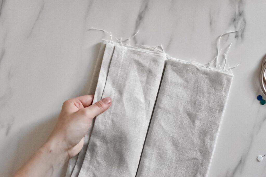 A hand holds a piece of beige fabric with frayed edges at the top, showing how to fold the fabric for a DIY linen ring sling. Colored pins and a sewing tool lie partially visible on the side, ready to transform the material into a baby carrier.