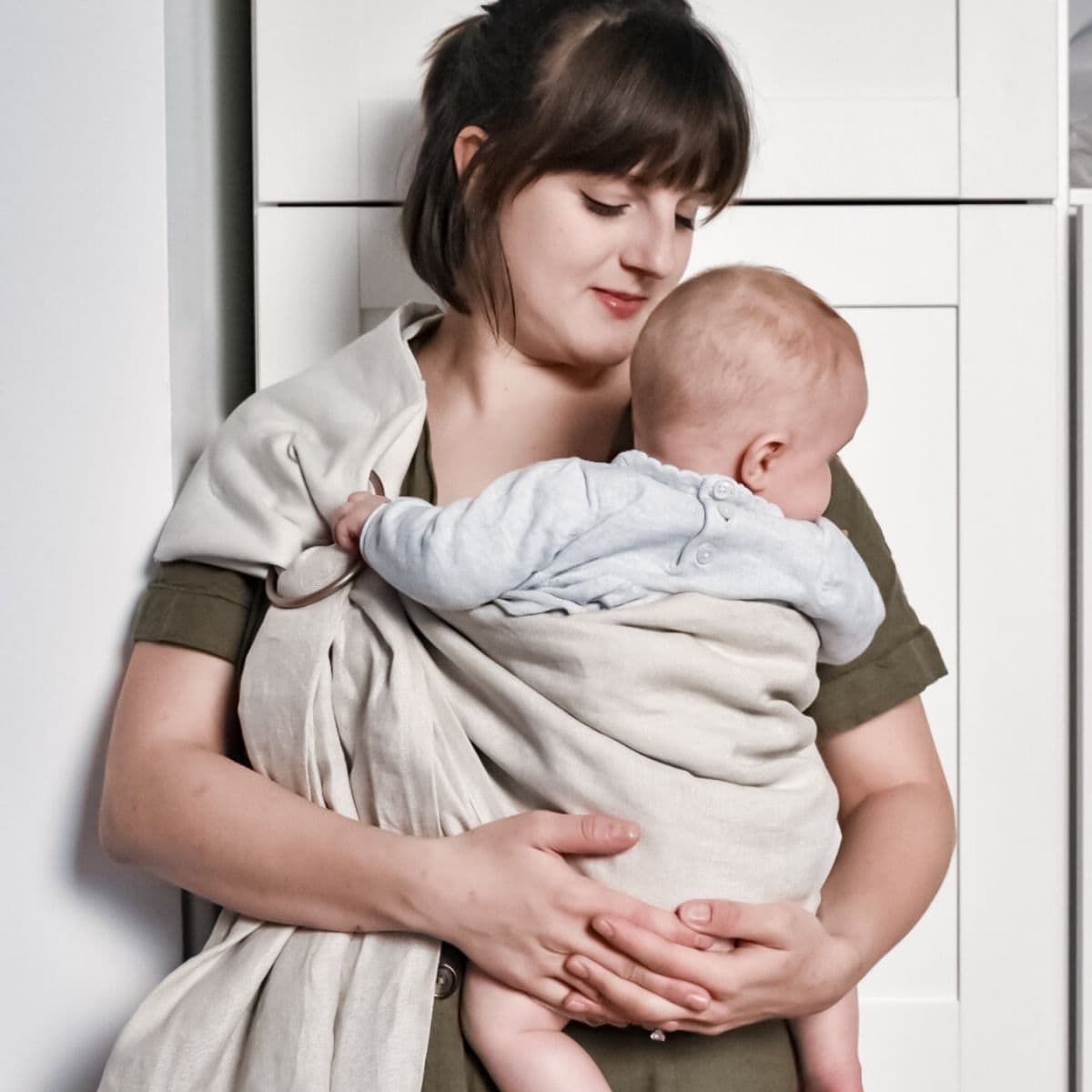 A woman with dark hair gently cradles a baby in a light-colored DIY linen ring sling against her chest while wearing an olive green top. She stands in front of white cabinets, and the baby, dressed in light gray, gazes away contentedly.