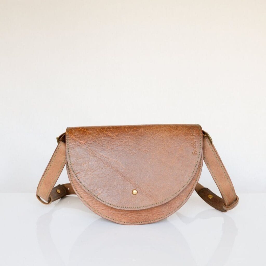 An eco-friendly gift idea, this brown leather crossbody bag features a semicircular flap and an adjustable strap, making it perfect for daily adventures. Displayed against a plain white background, its a stylish yet sustainable choice.