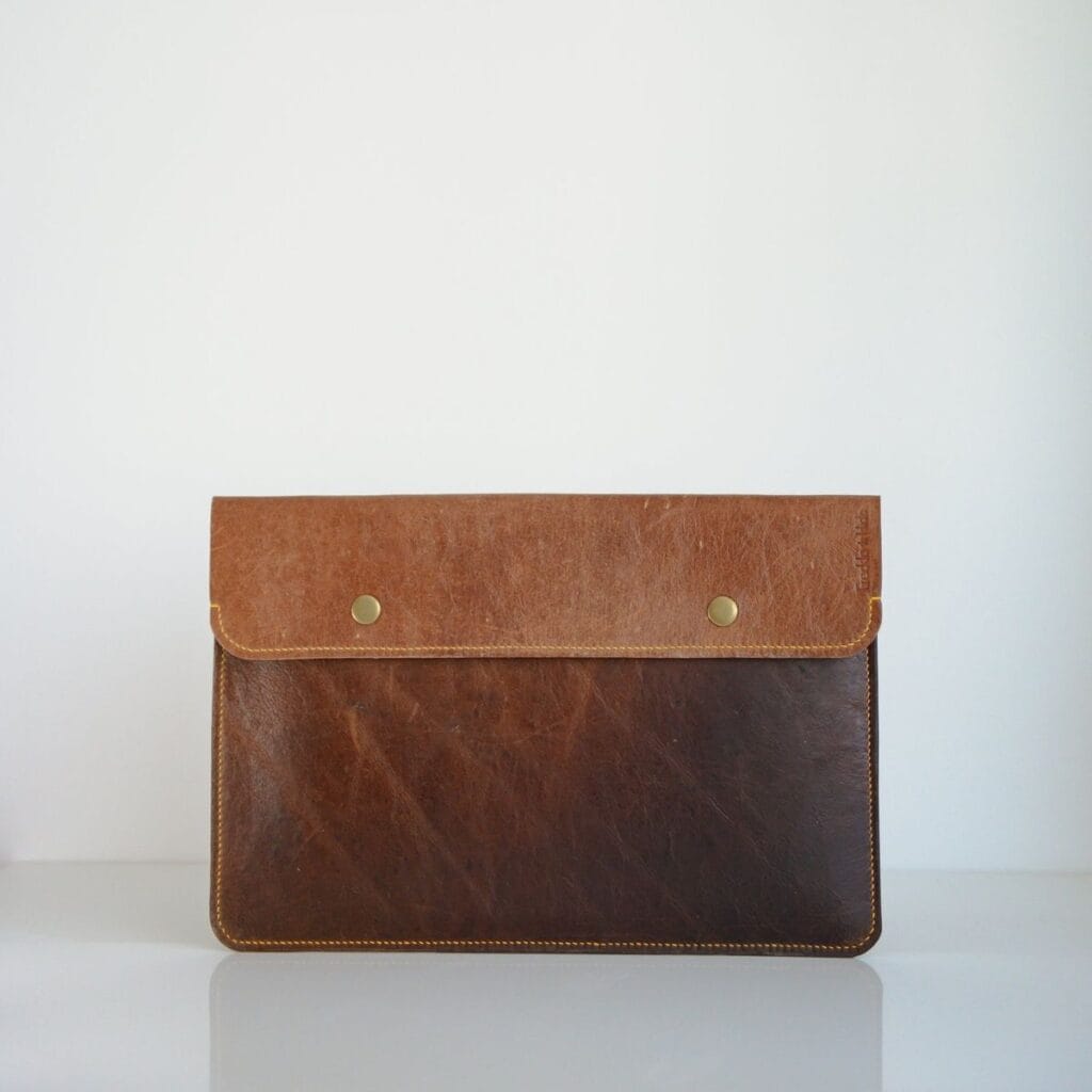 A brown leather laptop sleeve with a two-tone design. The top flap is a lighter brown, secured with brass buttons, while the lower part boasts a darker shade. It rests against a plain white background.
