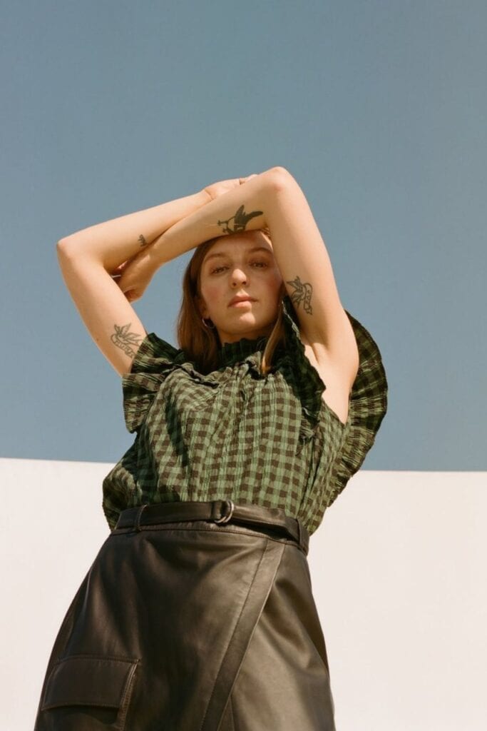 A woman with tattoos on her arms poses against a clear blue sky. Her green checkered top and black leather skirt complement the serene scene as she raises her arms above her head, gazing downward with a relaxed expression.