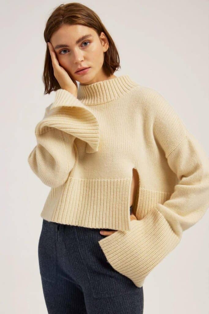 A person with shoulder-length brown hair is wearing a beige turtleneck sweater with flared sleeves and a side slit, showcasing an eco-friendly gift option. They pose elegantly with one hand on their face and the other on their hip, against a plain background.