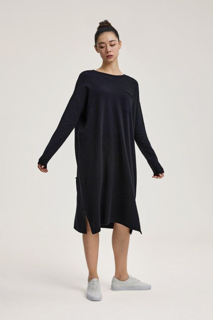 A person stands against a plain background, wearing a loose-fitting, long-sleeve black dress with a round neckline and side slit, paired with white sneakers. Their hair is styled in a bun and they exude relaxed elegance.