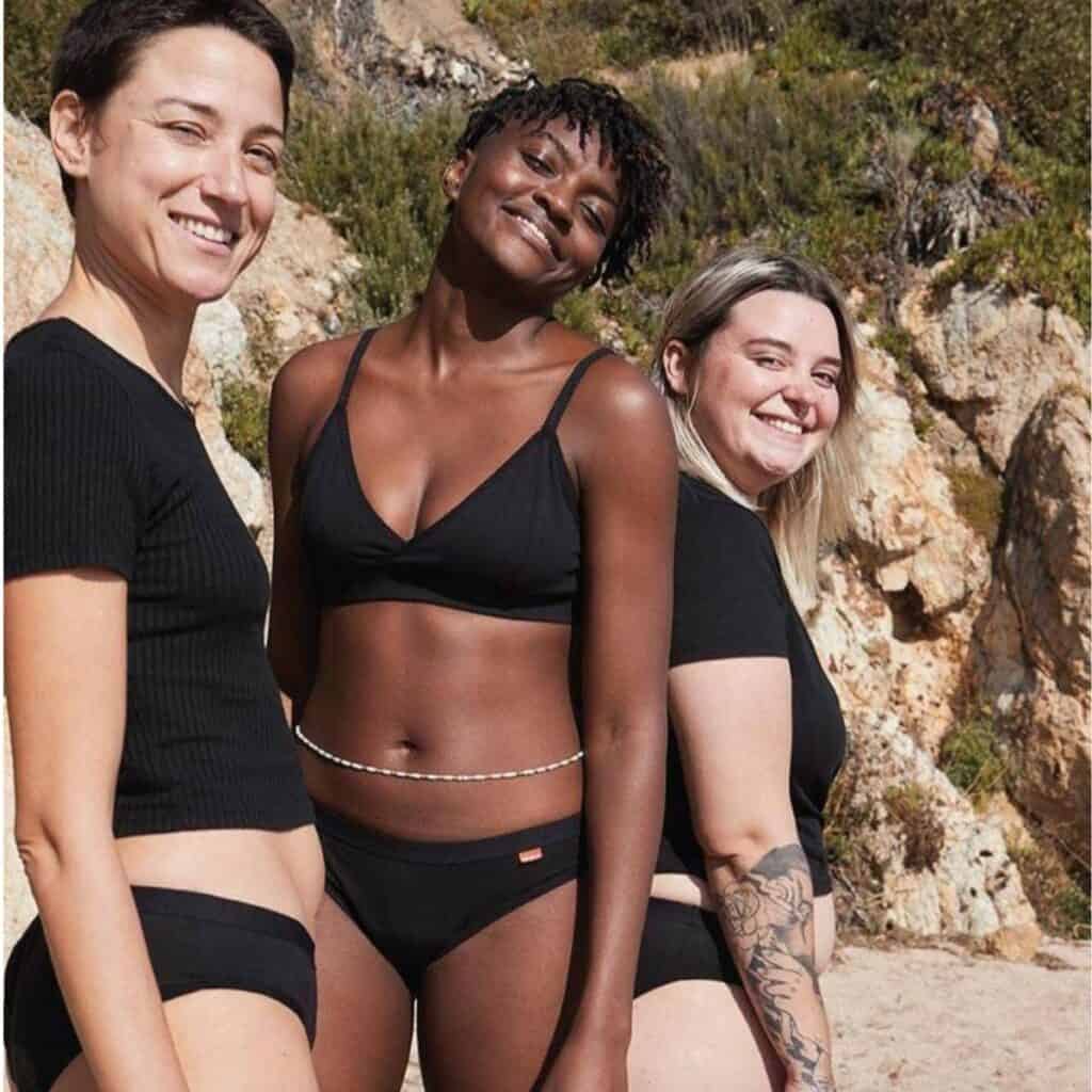 Three people stand confidently on a beach, smiling at the camera. They are wearing black period swimwear and are surrounded by rocky terrain and greenery, an eco-friendly backdrop.