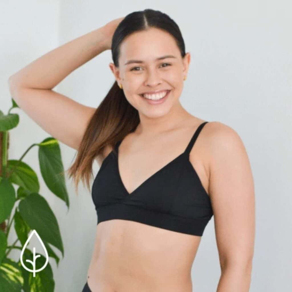 A person with long hair, smiling in a black bralette, stands before a lush green plant. A white leaf icon adorns the bottom left corner, hinting at eco-friendly gifts for her.