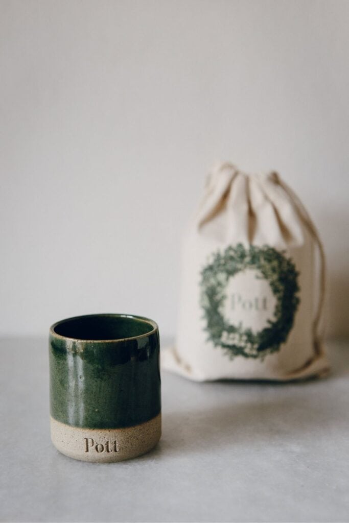 In the foreground sits a green ceramic candle pot with a beige base and Pott printed on it. Behind, a drawstring bag showcasing a leafy wreath design surrounds the Pott logo. This minimalistic setting makes for perfect eco-friendly gifts for her or him.
