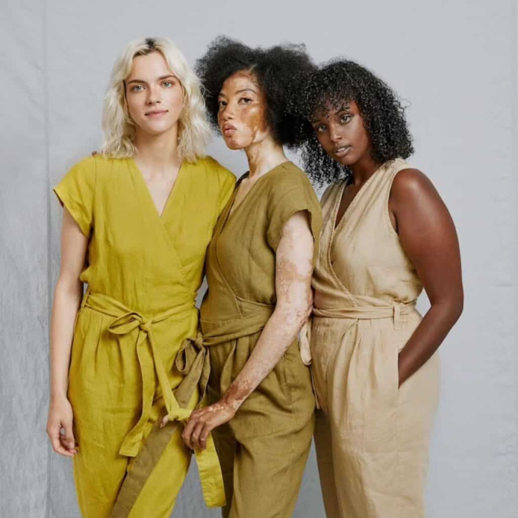 Three women wearing earthy-toned jumpsuits stand together against a plain backdrop, exuding confidence. The woman on the left has blonde hair, the middle woman showcases her vitiligo with pride, and the one on the right boasts curly hair. 