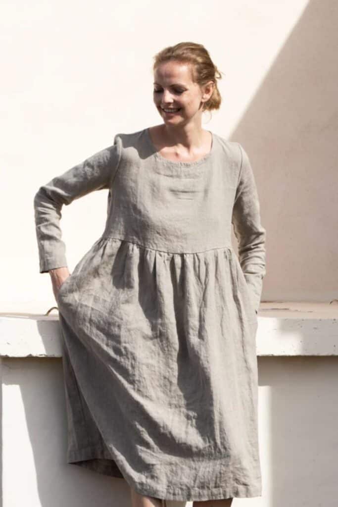 A person wearing a long-sleeved, loose-fitting gray dress stands outdoors, smiling and looking down. The eco-friendly dress boasts a gathered waistline. In the background, a light-colored wall enhances the serene moment.