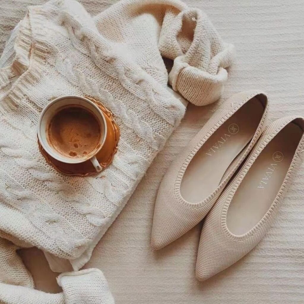 A cozy beige knit sweater lies next to a foamy cup of coffee and elegant beige pointed-toe flats, perfectly curated as eco-friendly gifts. Arranged on a soft, textured surface, they create a warm, inviting atmosphere ideal for thoughtful gifting.