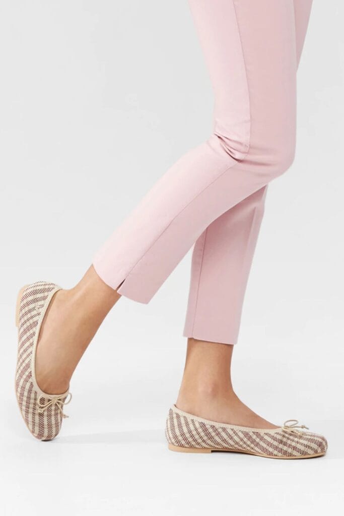 The person is shown partially, focusing on their lower legs in light pink pants and beige woven flat shoes with a bow detail—a perfect choice for eco-friendly gifts—against a plain white background.