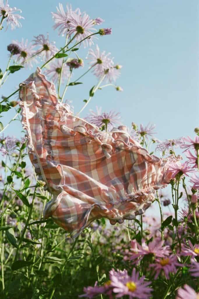 A pair of eco-friendly, pink and orange checkered frilled underwear sways gently among tall, blooming purple and white wildflowers under a clear blue sky. Her choice highlights the soft textures of the fabric and delicate petals in the sunlight.