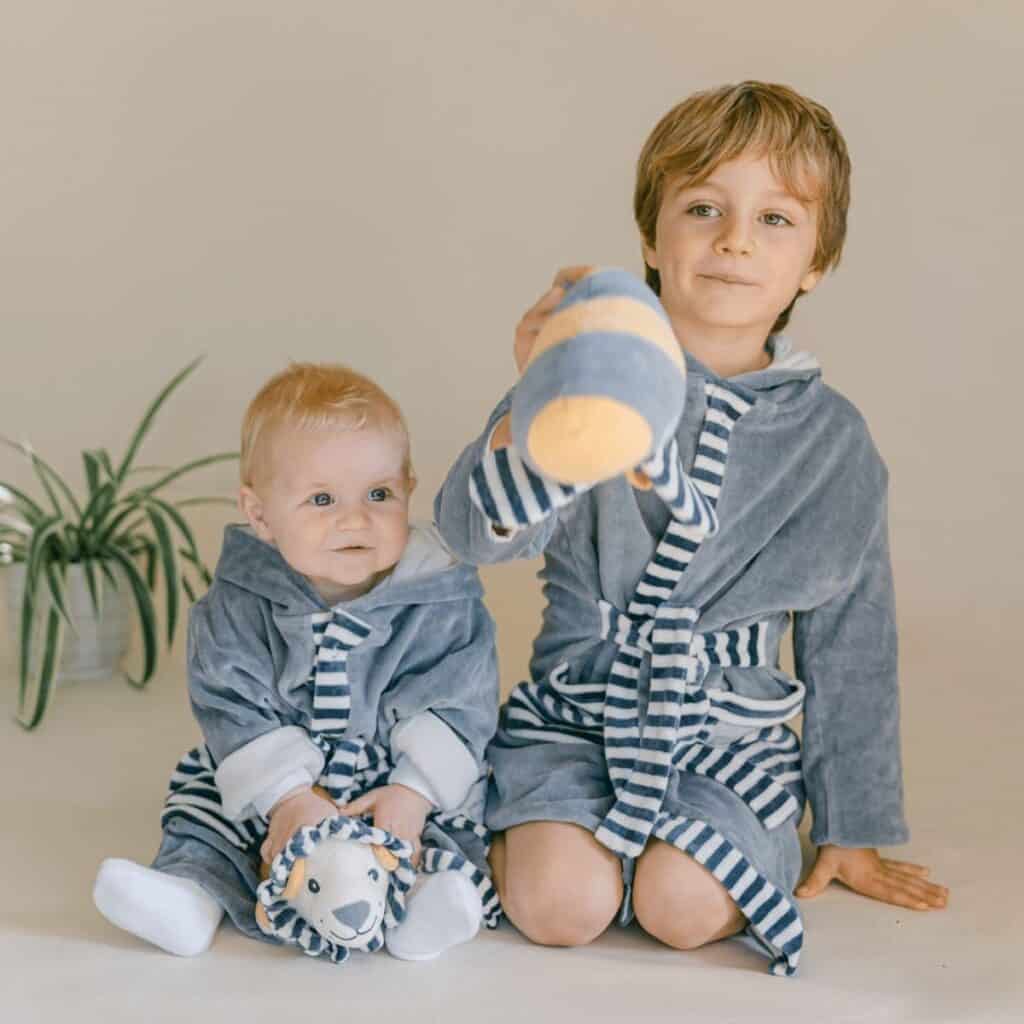 Two children sit together, wearing matching gray and striped robes. The older child holds up an eco-friendly toy with a striped pattern, while the younger one cuddles a plush zebra. A potted plant is visible in the background.