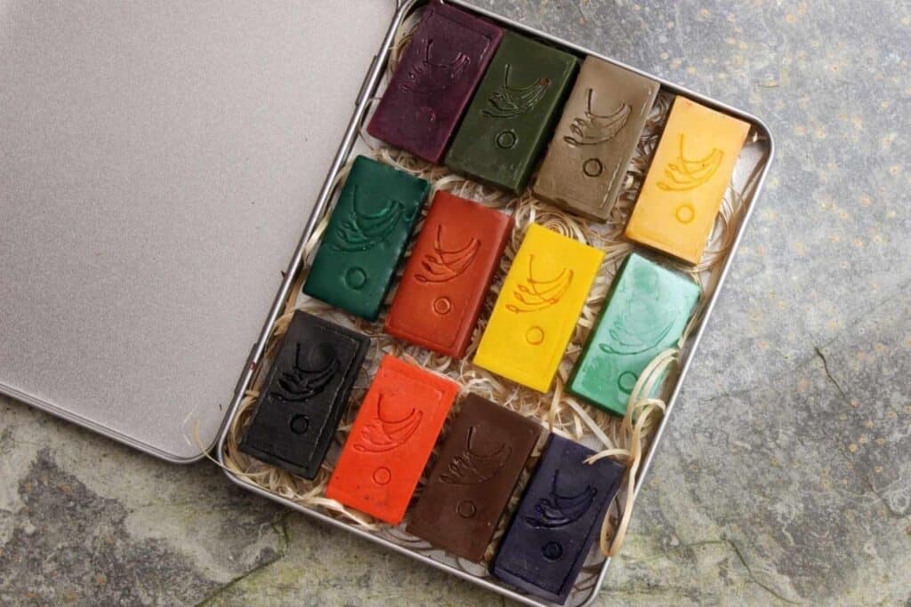 A metal tin holds twelve rectangular wax blocks in varying colors like green, brown, red, yellow, and blue. Each block is embossed with a leaf design and arranged neatly on a bed of shredded paper. Perfect as eco-friendly gifts for children, the open tin rests on a stone surface.