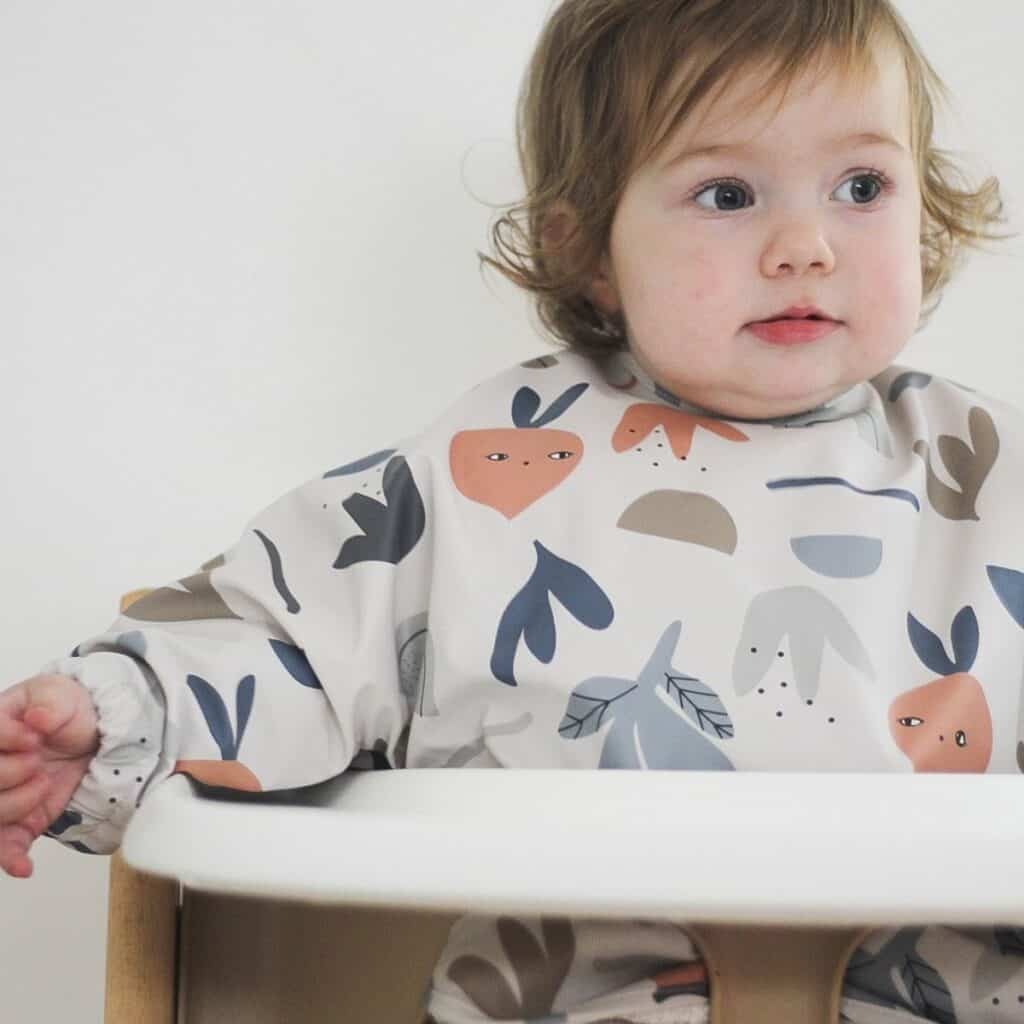 A toddler with light brown hair sits in a high chair, wearing a bib decorated with playful, colorful patterns of abstract shapes and faces. The child gazes to the side with a neutral expression. The plain, light-colored background highlights this serene moment.
