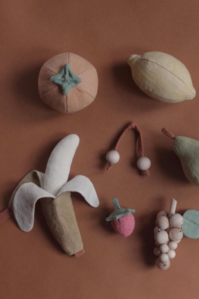A delightful collection of stuffed fabric toys in the shape of a banana, lemon, persimmon, strawberry, grape cluster, and cherries rests on a brown surface. Perfect as eco-friendly gifts for children and babies alike.