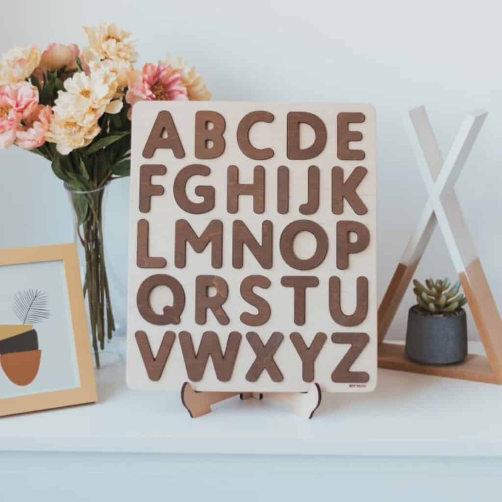A wooden alphabet board featuring cutout letters from A to Z is displayed on a stand. Perfect for children, its surrounded by pink and white flowers in a vase, a framed abstract art piece, and a succulent plant on a shelf.