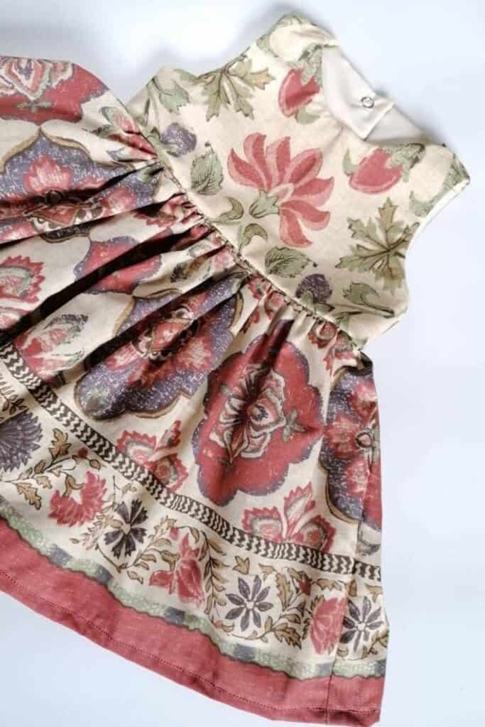 A sleeveless toddler dress with a floral pattern in red, green, and beige hues. Perfect for children, it features a full skirt and a button at the back. An eco-friendly gift choice, this charming dress is laid flat on a white background.