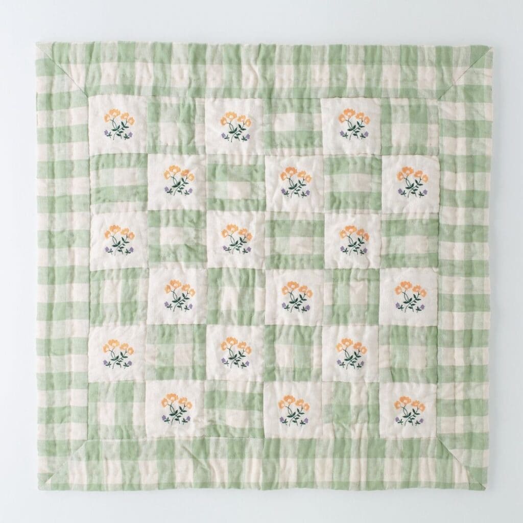 A square quilt with a green and white checkered pattern, featuring floral embroidery of orange flowers and green leaves. This charming quilt, perfect as an eco-friendly gift, boasts a symmetrical design neatly arranged on a light background.