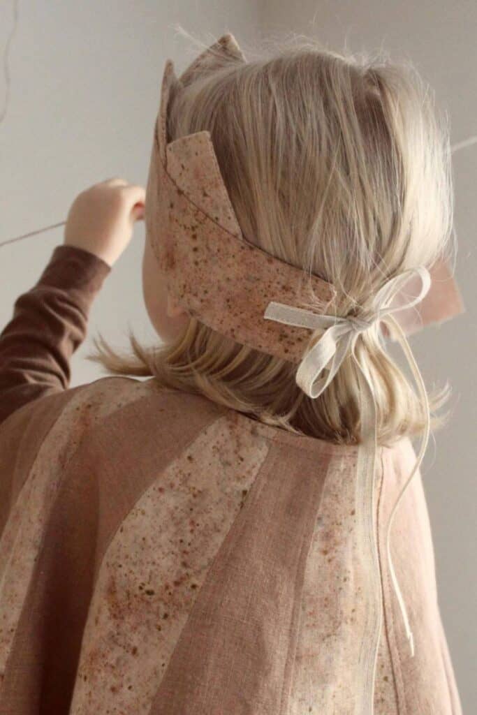 A child with light hair, seen from behind, wears a brown and beige costume with a matching headband. They hold a string in one hand in a softly-lit room.