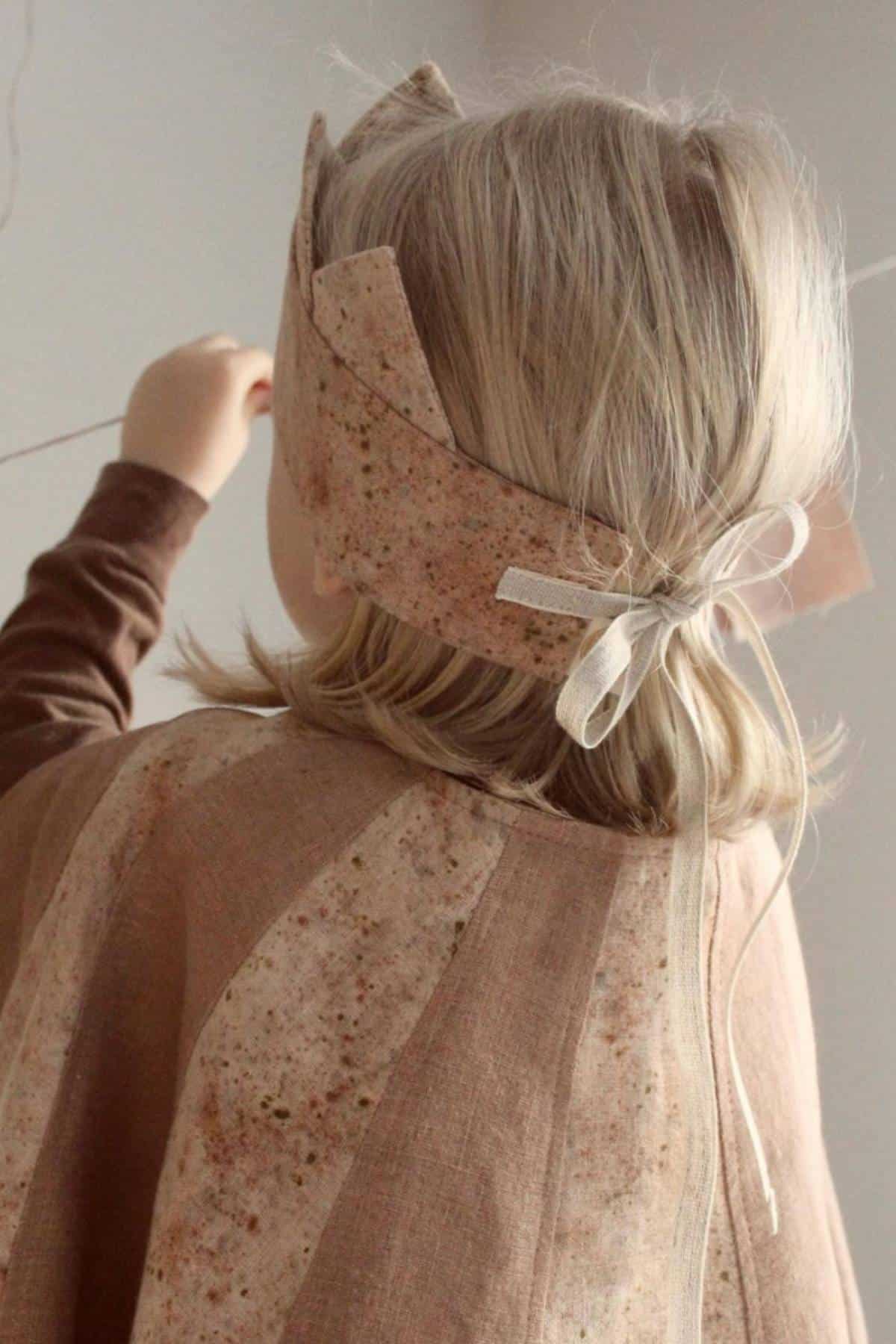 A child with shoulder-length blonde hair, seen from behind, stands indoors wearing a brown costume adorned with a headband tied with an eco-friendly ribbon.
