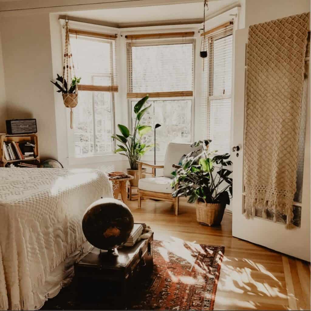 A cozy, sunlit bedroom showcases eco-friendly decor with a large window, wooden blinds, and potted plants. A white bed with a textured cover sits near a globe on a low table. A wicker chair with cushions by the window complements the woven tapestry on the door.