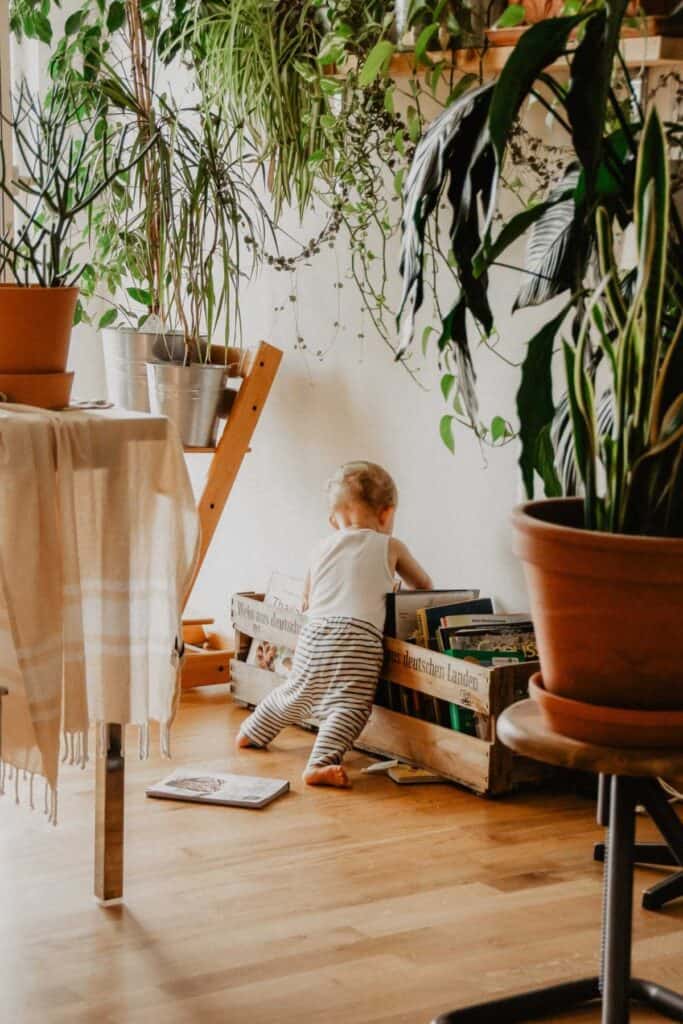 A toddler in a white tank top and striped pants stands on a wooden floor, surrounded by lush potted plants, creating an eco-friendly space. The child reaches into a wooden crate filled with books. A table with a cloth is partially visible on the left.