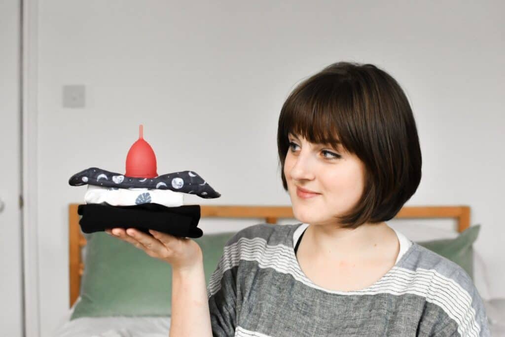 A person sits on a bed holding a stack of folded clothes topped with a red menstrual cup, embracing the eco-friendly menstruation movement. They have short brown hair and wear a gray striped shirt. Green pillows accent the background.