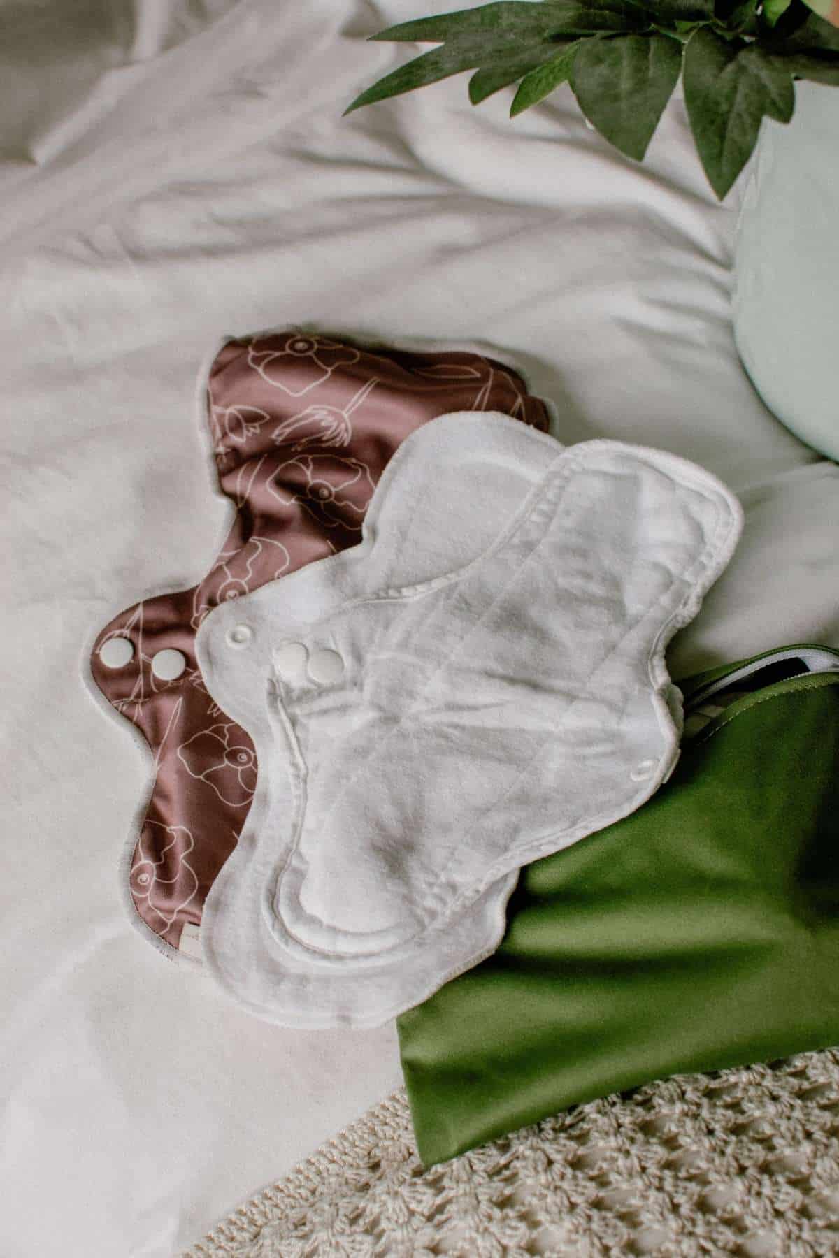 White and patterned cloth sanitary pads are neatly placed on a bed. A green bag and potted plant add a touch of nature to the eco-friendly menstruation scene in the background.