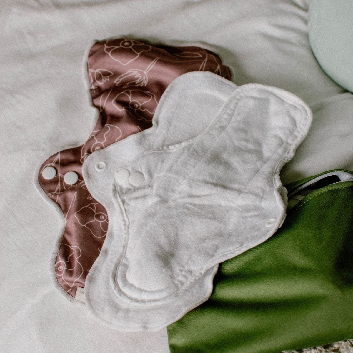 Two eco-friendly period pads are placed on a white surface. One cloth sanitary pad is white, and the other features a brown pattern. Both pads include snaps for secure fastening. A green fabric item is partially visible to the side.