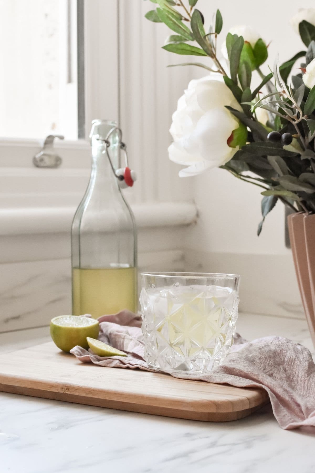 A glass of elderflower cordial sits on a wooden cutting board, a half-sliced lime, and small spoon beside it. In the background, theres a glass bottle and a potted plant with white flowers near a window.