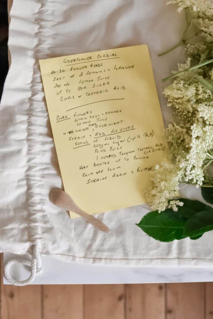 A handwritten elderflower cordial recipe graces a yellow paper on a white cloth. Freshly foraged elderflowers and a small wooden spoon accompany the scene, adding a rustic and natural touch.