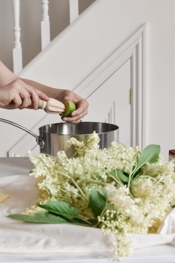 A person squeezes juice from a lime into a pot using a reamer. Fresh elderflowers lay on the table, hinting at a fragrant elderflower cordial in the works. A staircase banister is visible in the background, adding an inviting touch to this culinary scene.