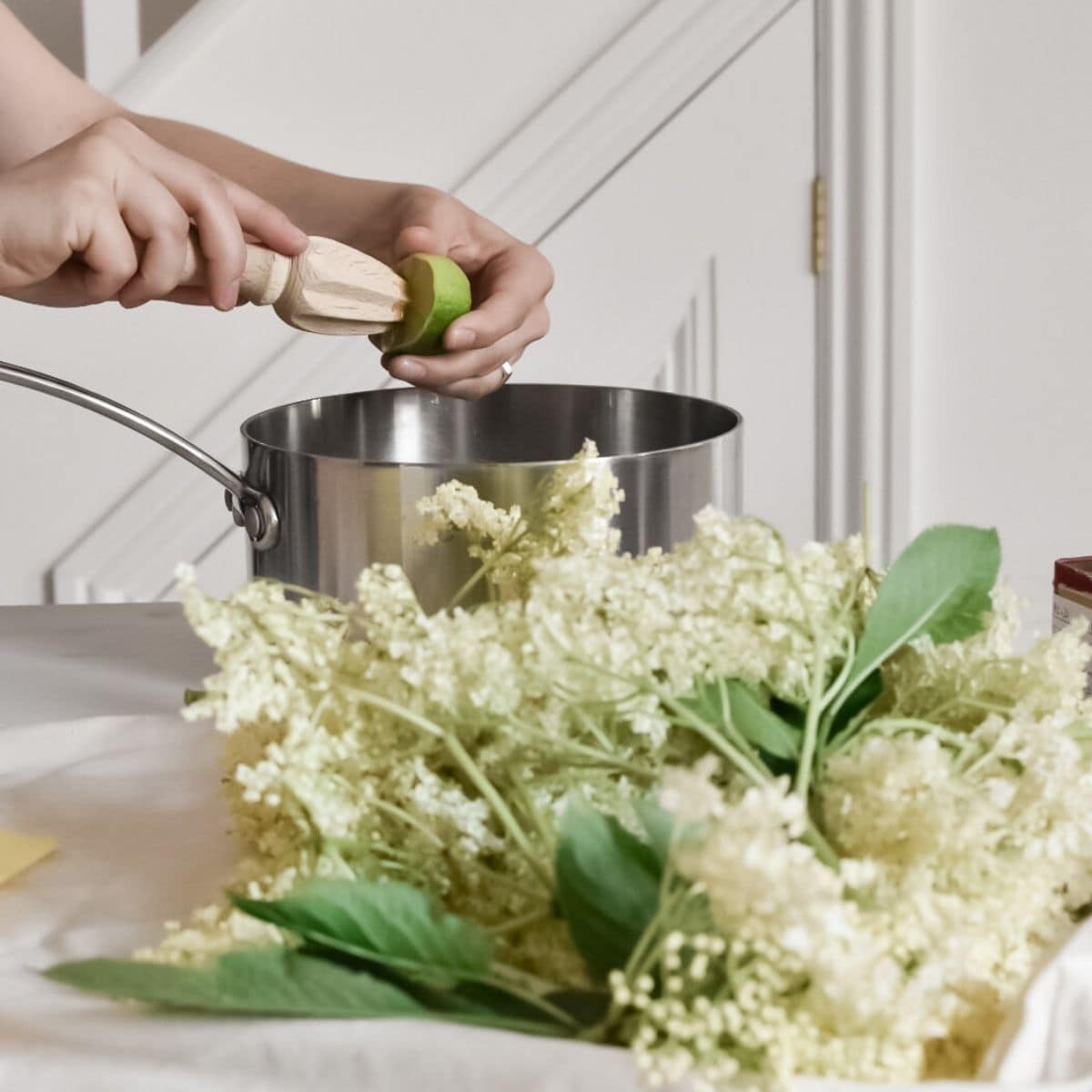 Hands using a wooden citrus reamer to juice a lime over a silver pot, crafting the perfect blend for elderflower cordial. In the foreground, freshly foraged white elderflowers with green leaves rest on the countertop. The background reveals a partial view of a white staircase.