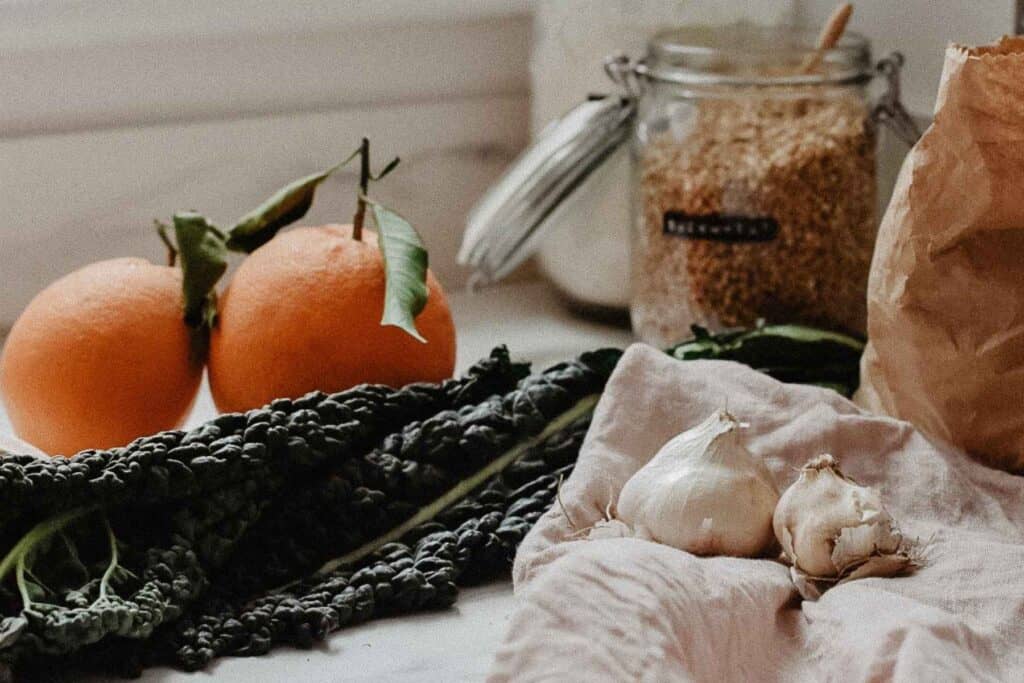 A rustic kitchen scene features two oranges with leaves, a bundle of dark leafy greens, garlic bulbs, a jar of grains, and a paper bag—all arranged on a light-colored cloth. This inviting setup inspires you to feed your family sustainably and create delicious meals affordably.