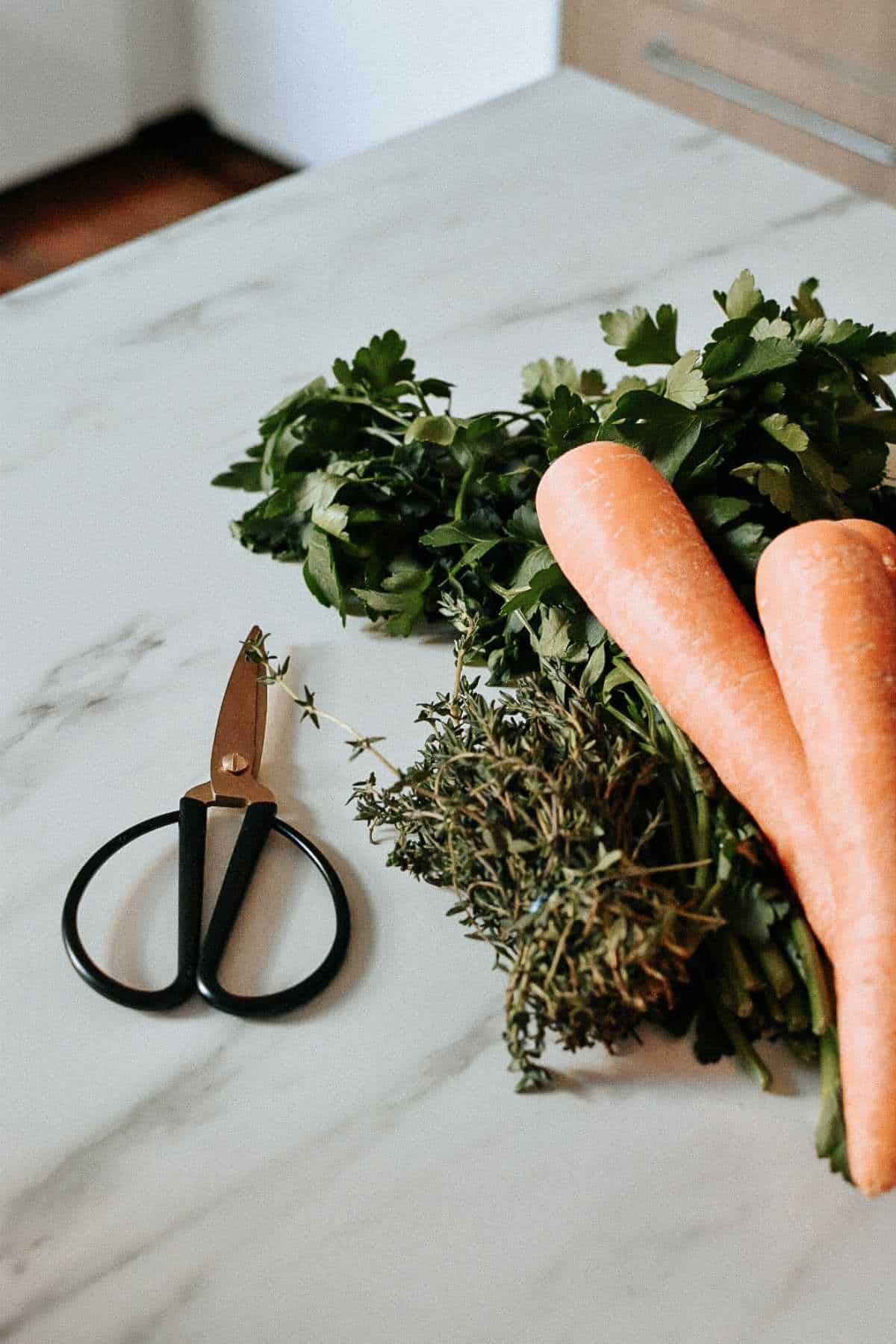 Carrots and fresh herbs, including parsley and thyme, rest on a marble countertop next to black-handled kitchen scissors.