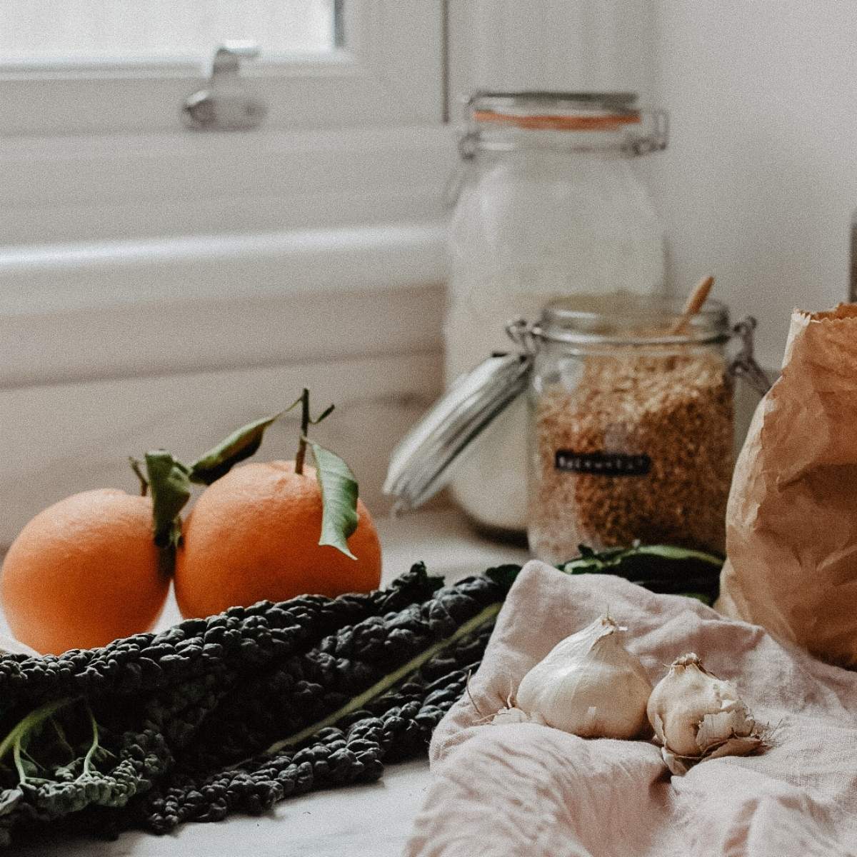A kitchen counter with two oranges, leafy greens, garlic bulbs, and open jars of flour and grains sits amidst a rustic window backdrop. A paper bag is partially visible, evoking a sustainably sourced vibe that feels both natural and family-focused.