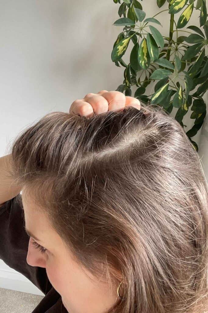 A person with brown hair is holding the top of their head to reveal a part in their hair. The hair density has significantly improved following the use of Feel Hair Support supplement.