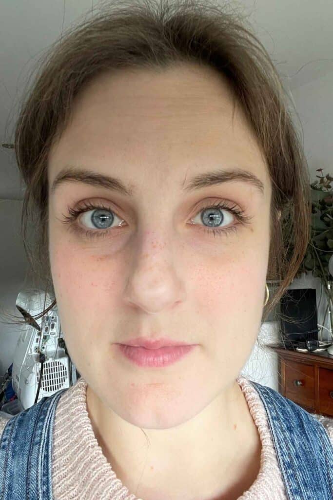 A person with blue eyes and brown hair looks directly at the camera. They are indoors, wearing a pink top with a denim jacket.