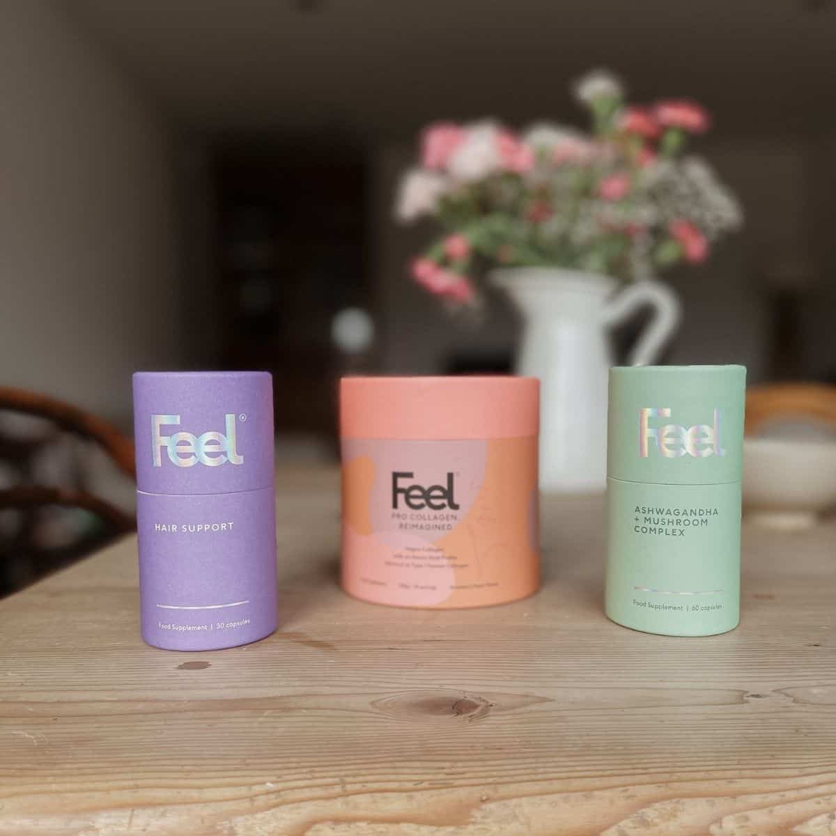 Three cylindrical containers on a wooden table, each labeled Feel. The purple one is for hair support, the coral one for collagen replenishment, and the green one for ashwagandha complex. A blurred vase with flowers is in the background.