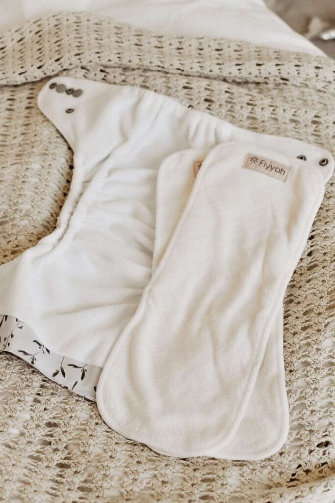 The Fiyyah Pokkit cloth diaper, featuring snap buttons and a subtle leafy edge pattern, rests on a textured woven surface. Its absorbent insert promises convenience and style, making it a top choice for those seeking an Econo Nappy solution.