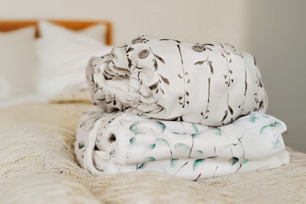 A stack of two patterned Fiyyah cloth nappies graces a bed with white and beige linens. The top nappy, a Fiyyah Pokkit nappy boasts a gray branch and leaf design, while the bottom, a Fiyyah Econo nappy, showcases a light blue leaf pattern.