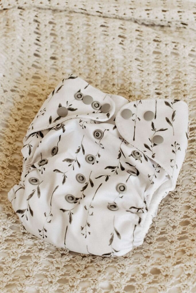 The Fiyyah Pokkit cloth diaper features a white background with a floral pattern, complemented by gray buttons and elastic edges. This Econo Nappy is beautifully displayed on a textured beige surface, offering both style and practicality.