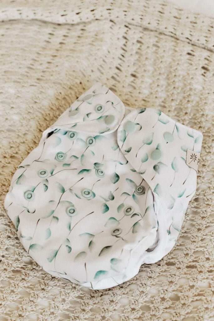 The Fiyyah Pokkit cloth diaper, with its floral pattern in soft greens and blues, rests elegantly on a textured beige surface, drawing attention from every parent eager for an Econo Nappy solution.