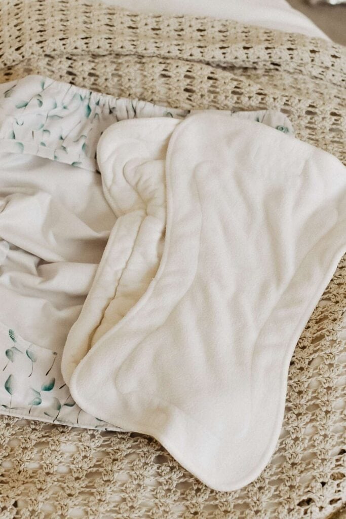 Cloth diapers and inserts, including the Econo Nappy, are laid out on a textured surface, featuring a floral-patterned cover and soft white liners.