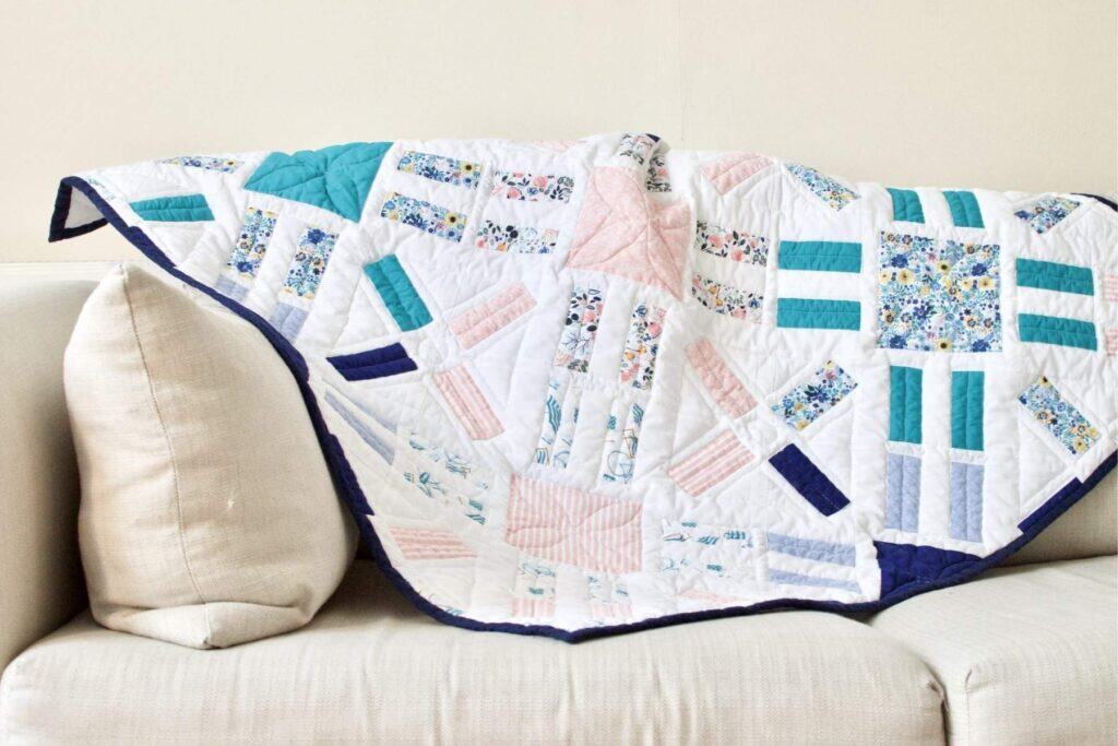 A colorful patchwork quilt featuring floral and geometric patterns is draped over the arm of a cream-colored sofa. With its pastel hues like pink, blue, and teal, this piece—crafted with expert quilting tips—creates a cozy and inviting atmosphere.