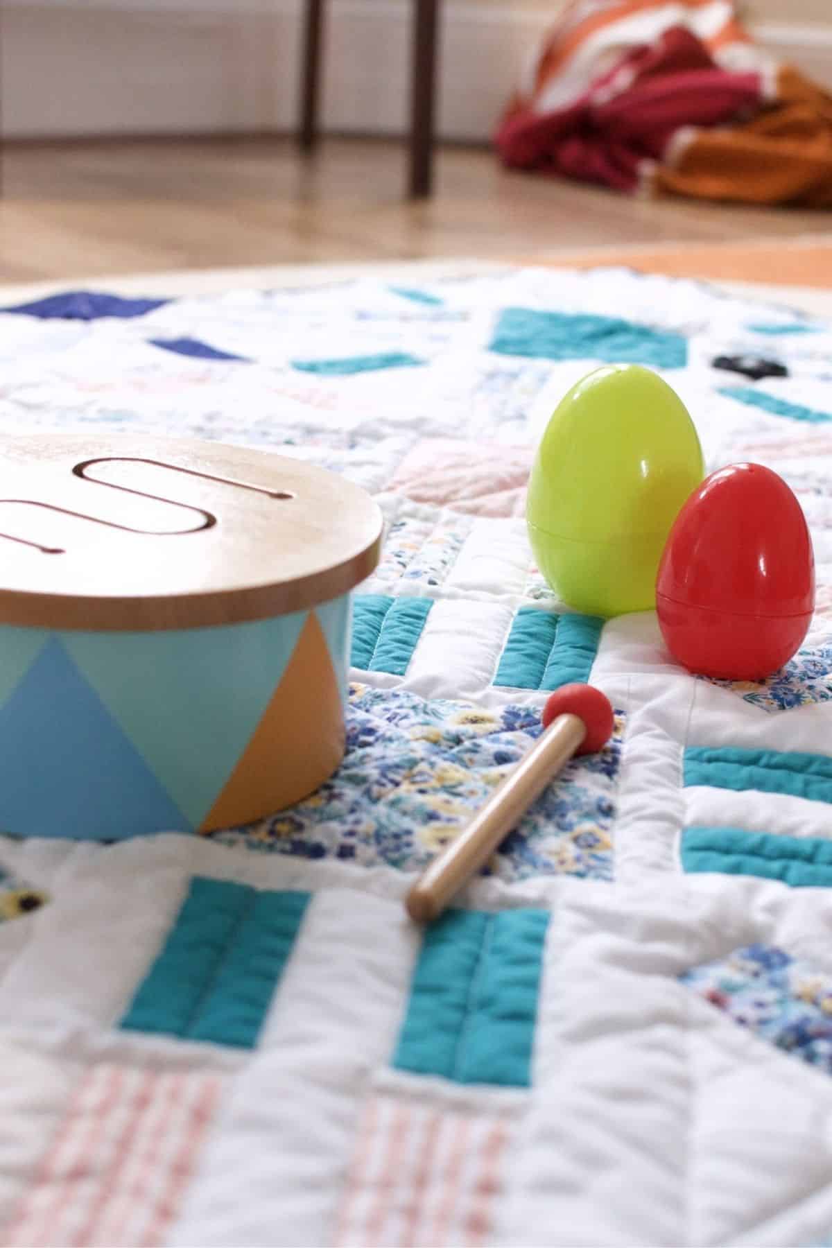 Childrens toy musical instruments, including a drum and colorful maracas, are placed on a patchwork quilt featuring blue, white, and floral patterns. 