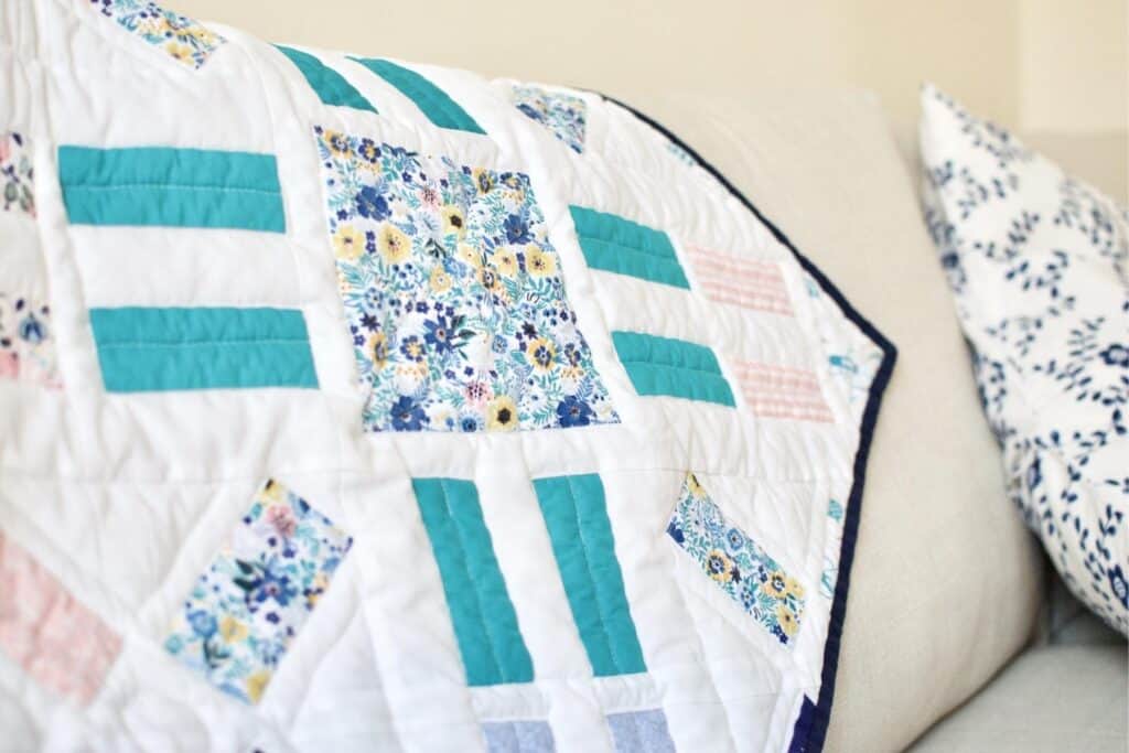A patchwork quilt with teal, floral, and patterned squares drapes over a beige couch, showcasing free motion quilting. In the background, a pillow adorned with a blue floral pattern adds a complementary touch.