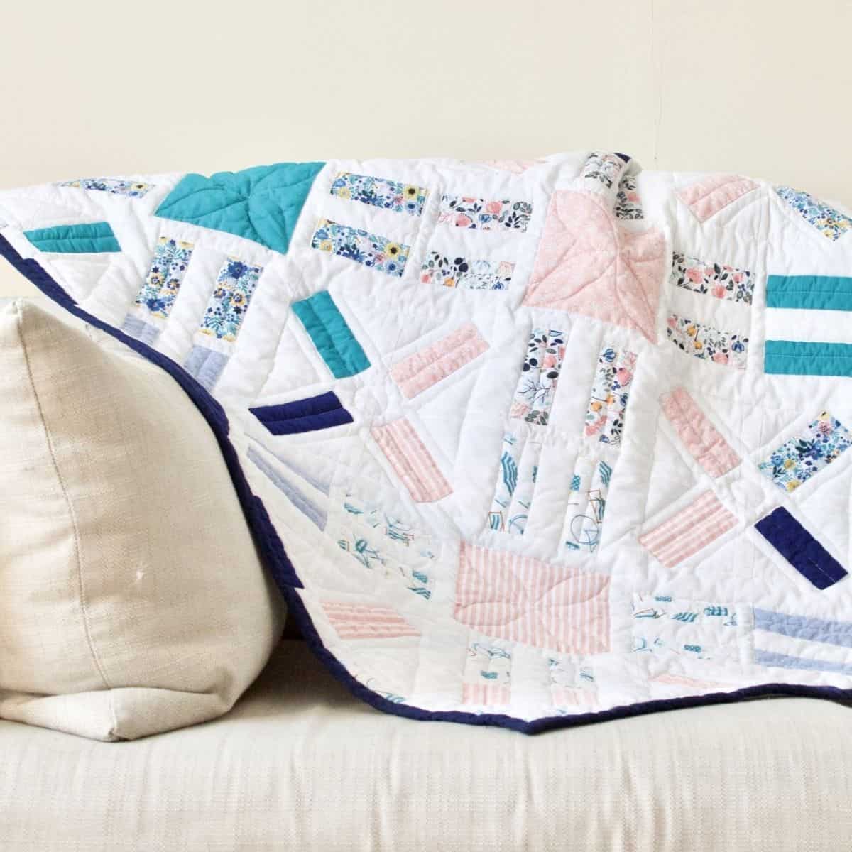 A colorful patchwork free motion quilt drapes over a light-colored sofa. It features floral and geometric patterns in pink, blue, and teal hues, framed by a dark blue border.