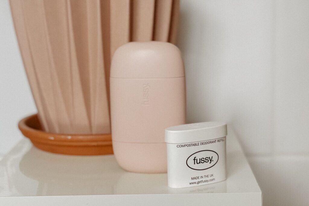 A minimalist, beige cylindrical deodorant container is placed next to a small white compostable deodorant refill tub labeled fussy. on a white surface. Theres a ridged terracotta pot in the background.